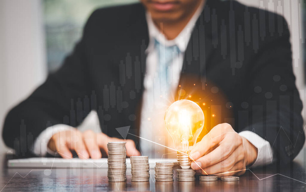 Рука бізнесмена тримає лампочку з бізнес графіками і стрілками і монетами стоїть на дерев'яному столі, збереження ідей та інвестиційного бюджету, концепція творчих ідей збереження грошей
,  - Фото, зображення