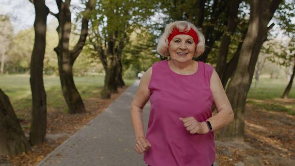 Starší stařena běží do parku a používá chytré hodinky. Žena běžec cvičení ven kardio venkovní - Fotografie, Obrázek