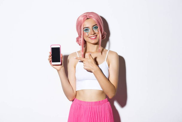 Πορτρέτο του χαρούμενου όμορφου γυναικείου μοντέλου σε ροζ glamour περούκα και φωτεινό μακιγιάζ, δείχνοντας το δάχτυλο στην οθόνη του κινητού τηλεφώνου, δείχνοντας εφαρμογή ή banner - Φωτογραφία, εικόνα