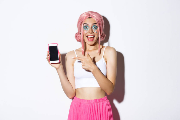 Portrait of excited beautiful girl in halloween costume, pink wig and bright makeup, pointing finger at mobile phone with amazed face, standing over white background - Photo, Image