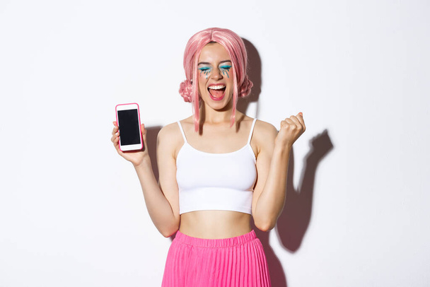 Immagine di una bella ragazza festaiola che si rallegra, festeggia le vacanze, mostra lo schermo dello smartphone e grida di gioia, indossa parrucca rosa e vestito di Halloween - Foto, immagini