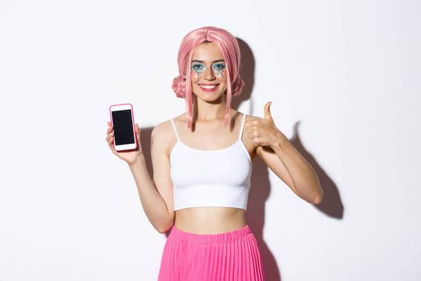 Εικόνα του ελκυστική νεαρή κοπέλα κόμμα σε ροζ περούκα, με φωτεινό μακιγιάζ, δείχνει αντίχειρες προς τα πάνω και οθόνη του κινητού τηλεφώνου, προτείνουμε app, στέκεται πάνω από το λευκό φόντο - Φωτογραφία, εικόνα