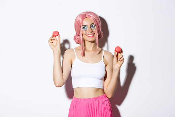 Mooie jonge vrouw in roze pruik tonen macarons en glimlachen, het eten van snoep, staande over witte achtergrond - Foto, afbeelding