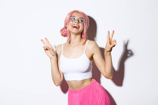 Portrait of beautiful smiling girl in pink wig, showing kawaii peace signs and laughing, wearing outfit for party, standing over white background - Photo, Image