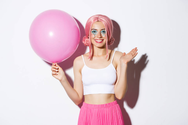Εικόνα της χαρούμενης νεαρής κοπέλας με ροζ περούκα και φωτεινό μακιγιάζ, κρατώντας μπαλόνι και χαιρετώντας το χέρι για να πει γεια, χαιρετώντας κάποιον στο πάρτι, γιορτάζοντας τις διακοπές, λευκό φόντο - Φωτογραφία, εικόνα