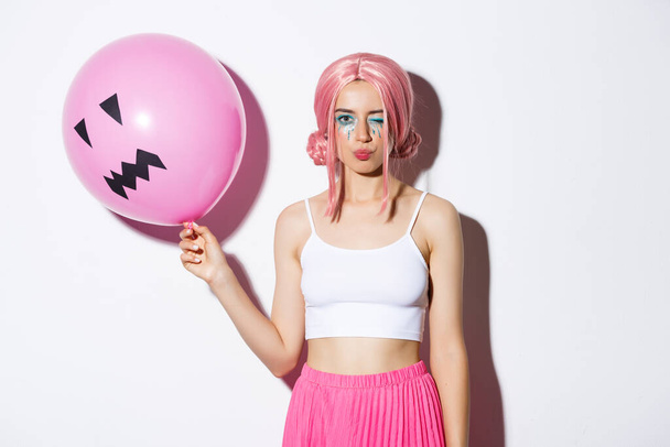 Εικόνα της αυθάδης όμορφη κοπέλα με ροζ περούκα και φωτεινό μακιγιάζ, κρατώντας μπαλόνι στο πάρτι, κλείσιμο του ματιού και αναζητούν coquettish, γιορτάζει τις διακοπές - Φωτογραφία, εικόνα