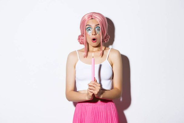 Porträt eines schockierten Mädchens, das zu Halloween Zaubersprüche zaubert, eine Kerze in der Hand hält und erstaunt schaut, im Kostüm mit rosa Perücke und grellem Make-up steht - Foto, Bild