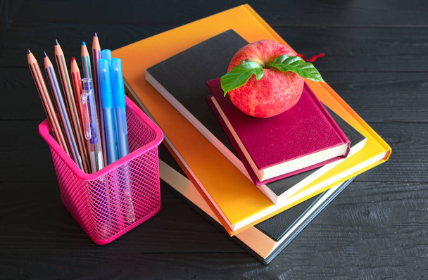 Stationery items on a black table. A stack of books and notebooks lie on a black background. Pencils and pens, stand in a pink jar for stationery. - Photo, Image