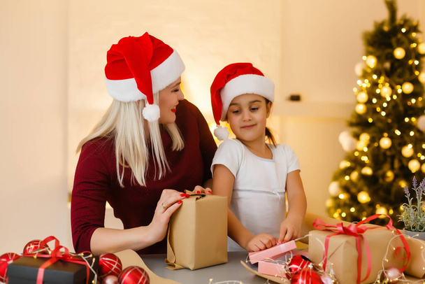 vacaciones, celebración, familia y concepto de personas - madre feliz y niña en sombreros de ayuda de santa con caja de regalo sobre la sala de estar y el fondo del árbol de Navidad - Foto, Imagen