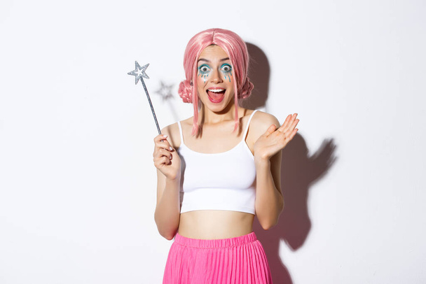 Retrato de una hermosa chica glamurosa celebrando Halloween con peluca rosa y disfraz de hadas, sosteniendo la magia y sonriendo feliz ante la cámara, de pie sobre un fondo blanco - Foto, Imagen