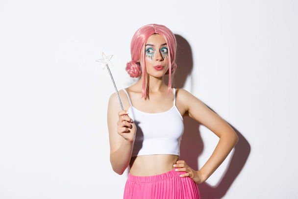 Image de belle femelle avec perruque rose et maquillage lumineux, tenant baguette magique, fée cosplay pour fête d'Halloween, debout sur fond blanc - Photo, image