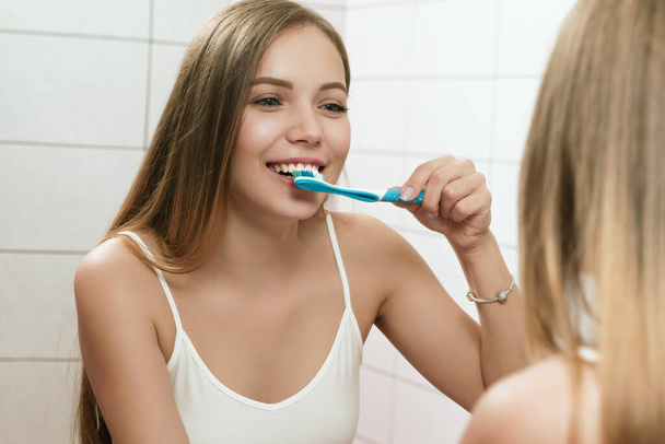 Молодая женщина чистит зубы в ванной. Отражение в зеркале - Фото, изображение