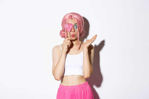 Portrait of attractive girl in pink wig holding cute candy over eye and pouting, wearing fairy costume on halloween, standing over white background - Fotó, kép