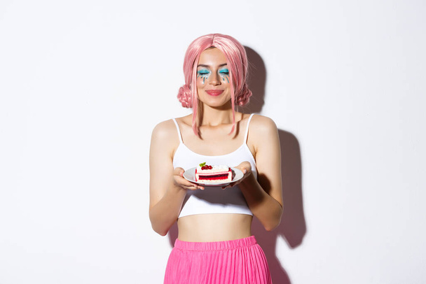 Afbeelding van mooi meisje in roze pruik sluiten ogen en ruiken smakelijke taart, vieren vakantie, staande over witte achtergrond - Foto, afbeelding
