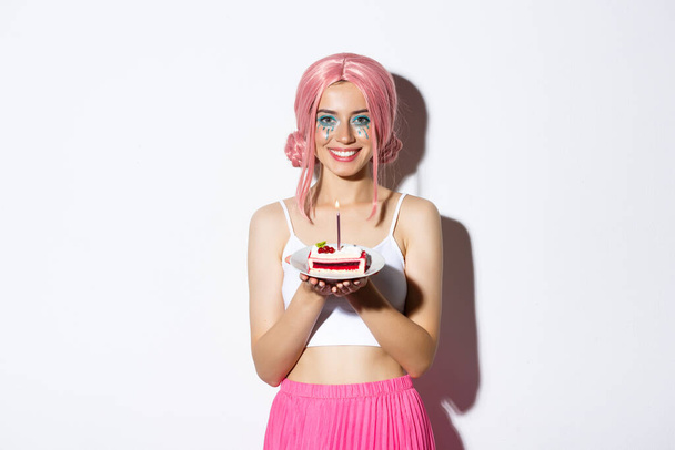 Immagine della bella festeggiata in parrucca rosa, con in mano la torta del b-day con la candela accesa, sorridente e desiderante, che celebra la festa su sfondo bianco - Foto, immagini