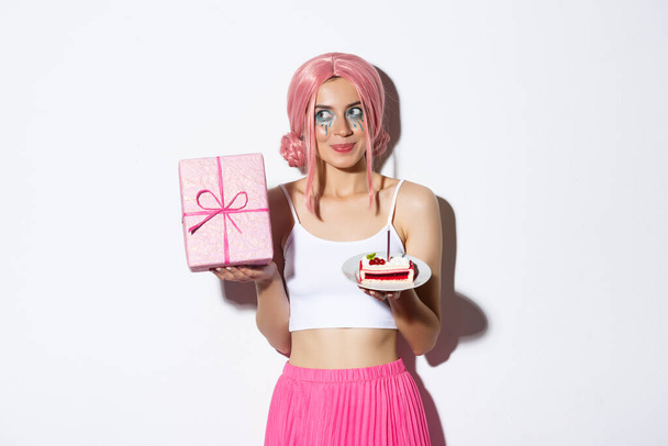 Immagine di eccitata ragazza carina in parrucca rosa, scuotendo scatola con regalo e vagare cosa all'interno, tenendo pezzo di torta di compleanno, celebrando b-day - Foto, immagini