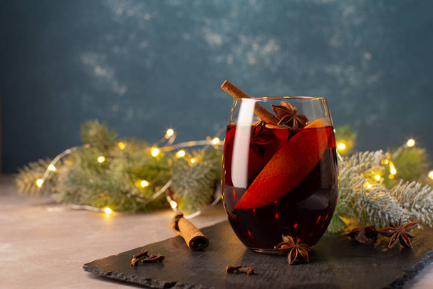 Christmas hot drink-Mulled wine with oranges,cinnamon,anise stars and cloves spice on festive Christmas background.Christmas cozy card with tree branches and christmas lights. - Zdjęcie, obraz