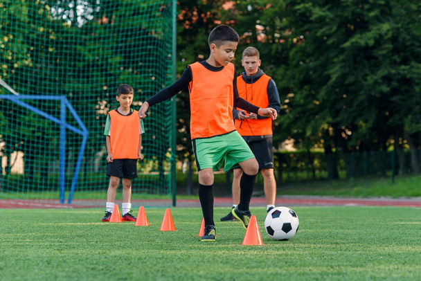 Wytrwały nastolatek w odzieży sportowej trenuje piłkę nożną na boisku do piłki nożnej i uczy się okrążać piłkę między stożkami treningowymi. - Zdjęcie, obraz