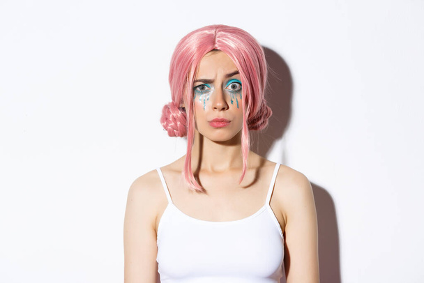 Close-up of confused girl in pink wig, raising eyebrow with suspicious expression, standing over white background - Photo, Image