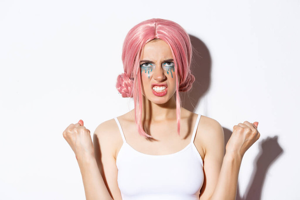 Lähikuva vihainen ahdistunut tyttö vaaleanpunainen peruukki, puristamalla nyrkit ja katselee ylös vihaa, seisoo valkoisella taustalla - Valokuva, kuva