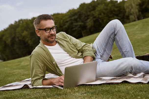 Arbeiten in der Natur. Junger Mann mit Brille liegt auf grünem Gras im Park und benutzt Laptop - Foto, Bild