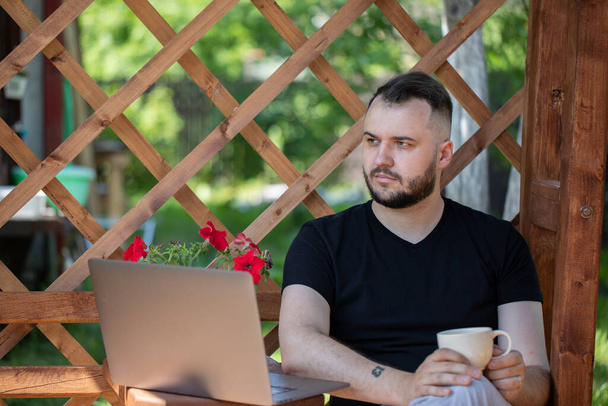 Beau jeune homme buvant du café fonctionne avec ordinateur portable à l'extérieur sur pergola en bois - Photo, image