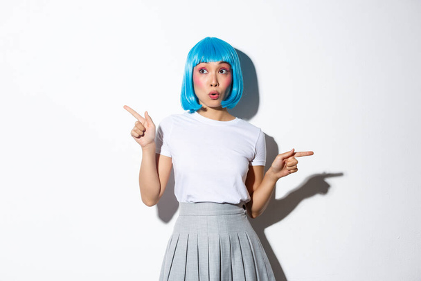 Obraz zdumionej azjatyckiej dziewczyny w niebieskiej peruce zwrócić uwagę na reklamy, wskazując palce bokiem i patrząc zdumiony, stojąc nad białym tle - Zdjęcie, obraz