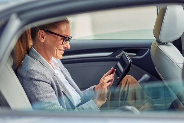 Filet de surf sur le chemin du bureau, vue latérale de belle femme d'affaires heureuse assise sur le siège arrière dans la voiture, en utilisant une tablette numérique et souriant - Photo, image
