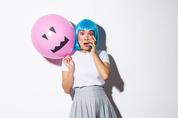 Retrato de assustado asiático menina no halloween traje e azul peruca, ofegante e olhando emboscado, segurando balão com assustador rosto - Foto, Imagem