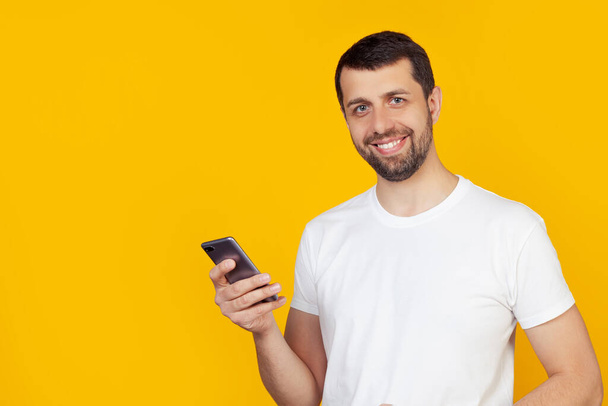 Young man with a beard in a white t-shirt is using a smartphone with a happy face standing and smiling with a confident smile showing teeth. Stands on isolated yellow background. - Foto, afbeelding