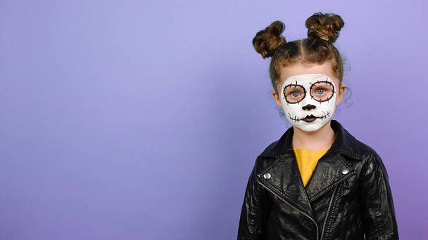 Portrait of child girl looking at camera, wears black leather jacket, isolated on purple studio background with copy space. Little girl wears frightening makeup. Kid prepares for mexican day of dead - Fotoğraf, Görsel