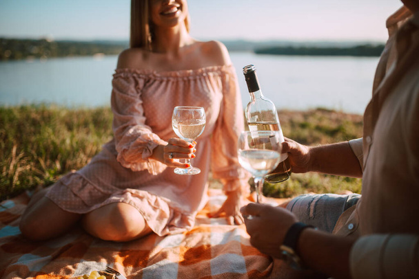 Close - up Urocza blondynka w pastelowej różowej sukience i mężczyzna pijący białe wino na plaży o zachodzie słońca. Kochająca się para świętuje i cieszy się piknikiem nad brzegiem rzeki - Zdjęcie, obraz