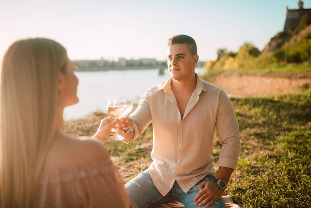 パステルピンクのドレスを着た可愛いブロンドの女性と、日没時にビーチで白ワインを飲む白人男性。川のほとりでピクニックをお祝いして楽しむ素敵なカップル - 写真・画像