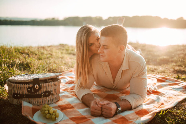 Un retrato de un hombre caucásico y una mujer rubia acostados en una manta en un picnic junto al río. Romántico feliz pareja amorosa en la puesta del sol - Foto, imagen