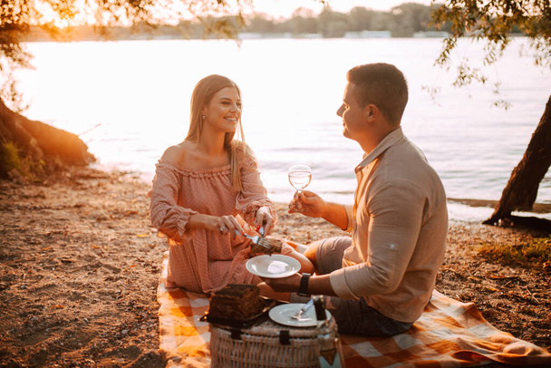 A beautiful caucasian woman in a pastel pink dress and a man are sitting on the beach and eating a cake. A loving couple celebrating a birthday on the river bank - Фото, изображение
