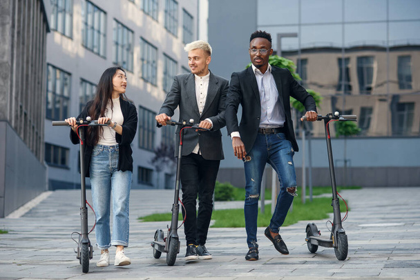 Confident happy multiracial office colleagues discussing business project go near office building with electric scooters - Valokuva, kuva