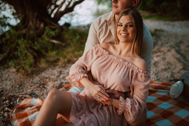 Un'attraente donna caucasica bionda giace tra le braccia di un uomo sulla spiaggia. Una coppia amorevole su un picnic vicino al fiume - Foto, immagini