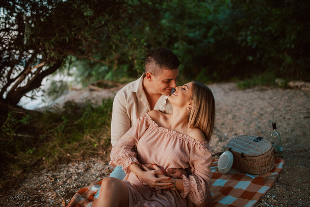 Una mujer atractiva con un vestido rosa pastel y un hombre besándose en una manta en la playa. Una pareja amorosa en un picnic junto al río - Foto, imagen