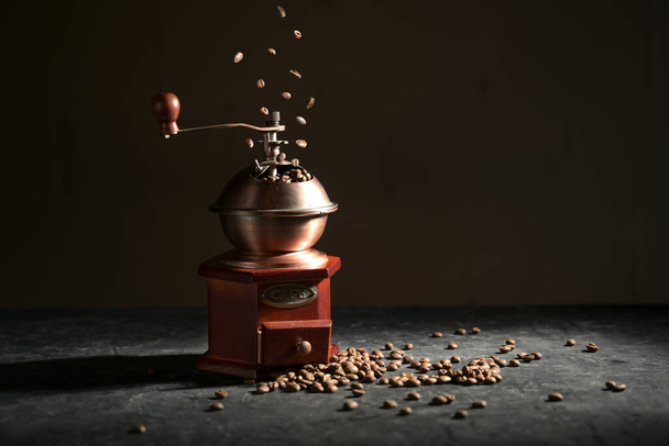 Wooden vintage coffee grinder on a slate plate, falling and lying roasted beans against a dark background with copy space, selected focus  - Foto, afbeelding