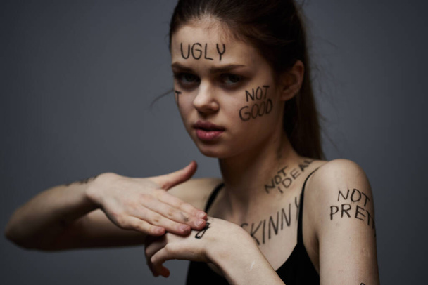 beleidigte Frau mit wütenden Schriftzügen auf ihrem Körper berührt sich mit den Händen auf grauem Grund  - Foto, Bild
