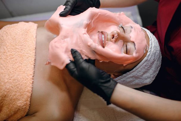 Брюнетка проходит процедуры в спа-салоне, розовая маска, нанесенная толстым слоем, снимается мастером в черных перчатках, очищает кожу от грязи, контролирует чистоту лица - Фото, изображение