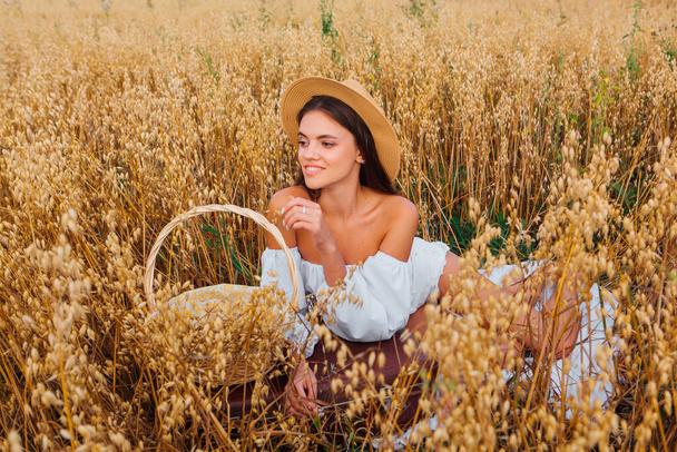 Rural Countryside Scene. Young beautiful woman with long hair dressed in white blouse and straw hat laying at golden oat field holding basket with ears of oats. - Foto, immagini