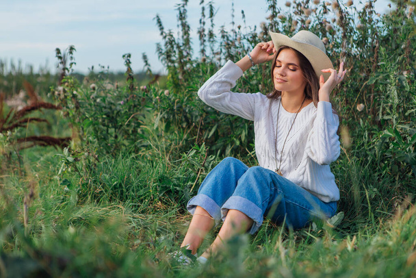Young beautiful brunette woman dressed in a white sweater, jeans and cowboy straw hat sitting on green grass, smiling and laughing during sunset - Foto, afbeelding