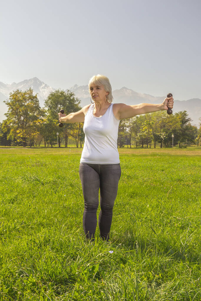 Elderly woman is engaged in fitness with a dumbbell in isolation mode outdoors in a park against a background of mountains on a sunny day - Zdjęcie, obraz
