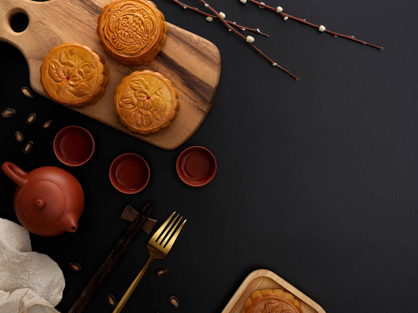 Top view of dark modern table with tea set, moon cakes on wooden tray, fork, decoration and copy space. Chinese character on the moon cake represent "five kernel and roast pork" in English  - Фото, изображение