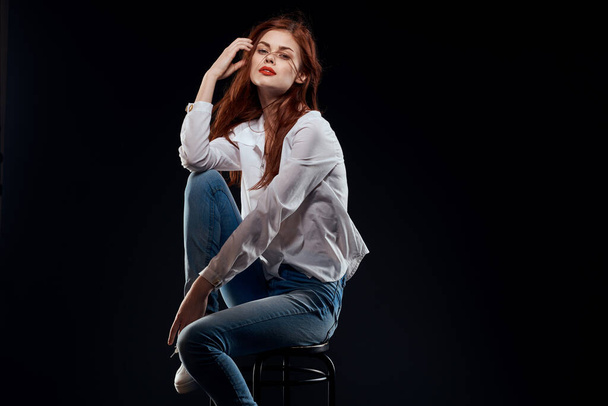 pretty woman sitting on a chair posing white shirt jeans long hair red lips dark background - Photo, Image