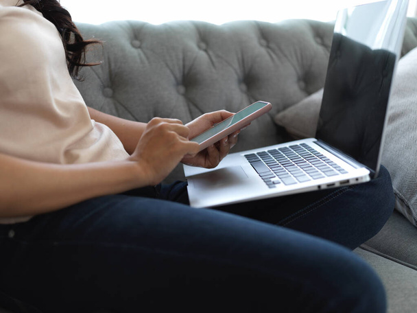 Vista lateral de la mujer usando el teléfono inteligente mientras trabaja con el ordenador portátil en su regazo en la sala de estar  - Foto, Imagen