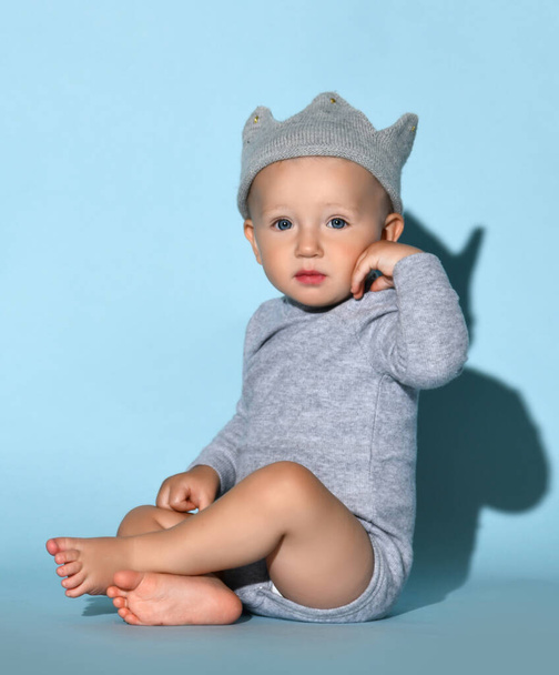 Toddler one-year-old baby boy in diaper and grey one-piece bodysuit with long sleeves and funny hat sits side to camera - 写真・画像