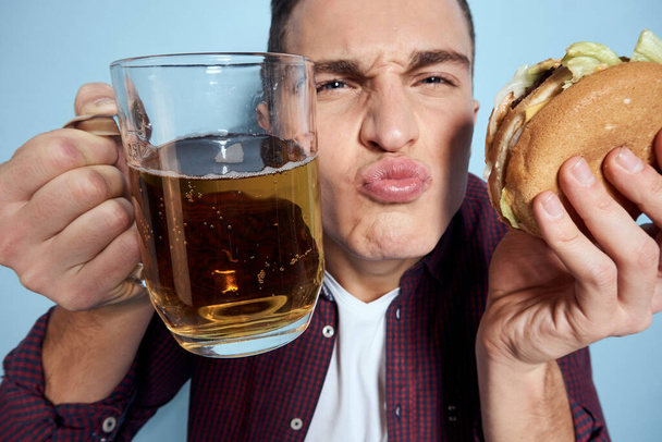 allegro uomo ubriaco con boccale di birra e hamburger in mano dieta cibo stile di vita sfondo blu - Foto, immagini