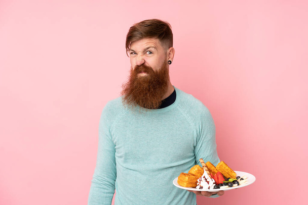Redhead man with long beard holding waffles over isolated pink background making doubts gesture while lifting the shoulders - Foto, Imagem
